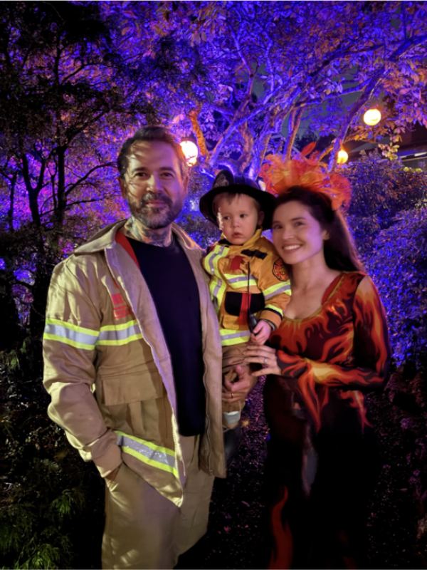 A family in costume — a firefighter, a small child dressed to match, and a figure in flame-coloured robes — under trees lit purple.