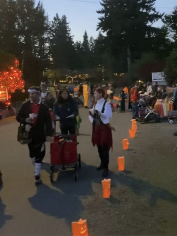 Neighbours in costume walking the closed block at dusk past rows of glowing paper lanterns.