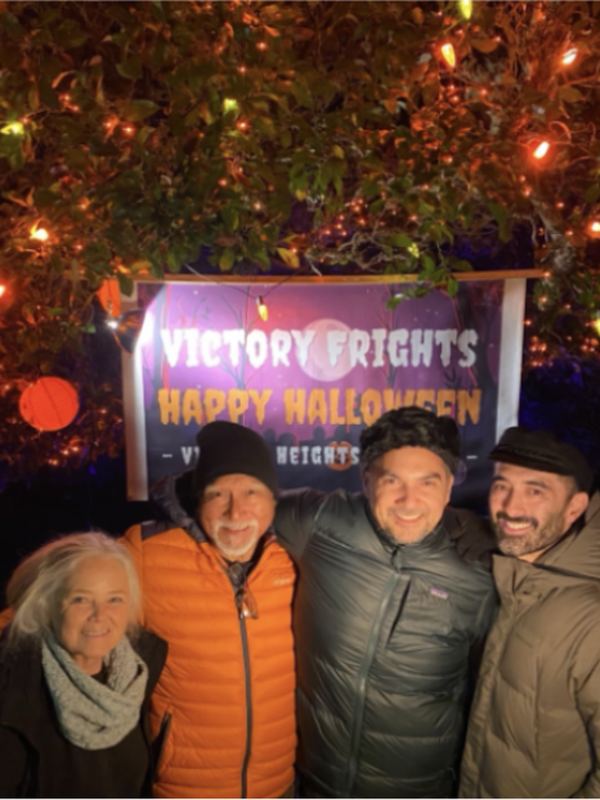 Four neighbours smiling under the Victory Frights Happy Halloween banner, surrounded by orange lights.